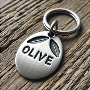 Könnte beinhalten: Silberne ovale Hundemarke mit gebürsteter Oberfläche und dem Namen "OLIVE" in schwarzen Lettern graviert. Die Marke hat zwei Olivenzweige oben.