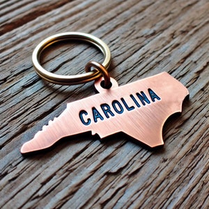 May include: A copper-colored keychain shaped like the state of North Carolina with the word "CAROLINA" engraved on it. The keychain is attached to a silver key ring.