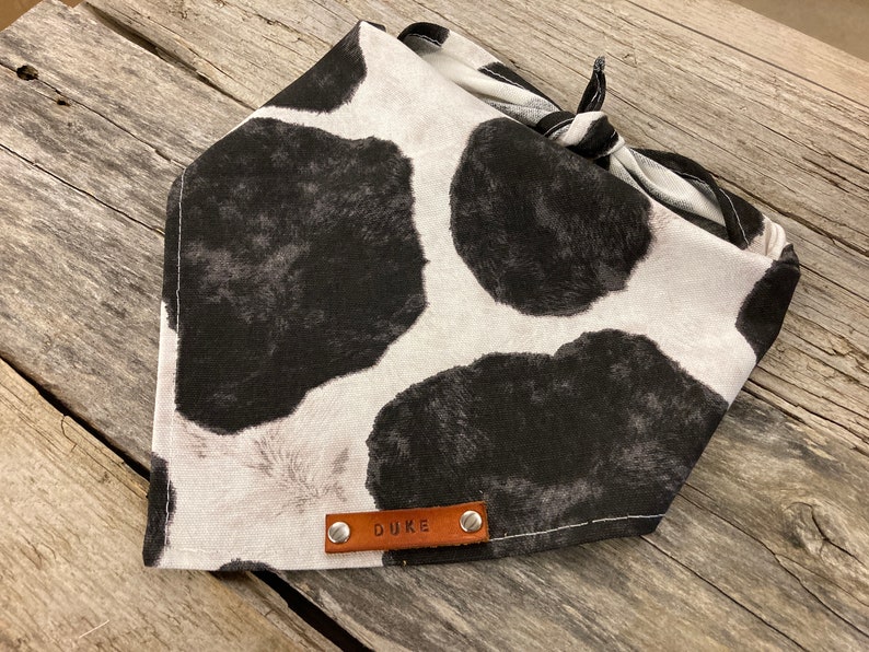 Peut inclure: Un bandana blanc et noir &agrave; imprim&eacute; vache avec une &eacute;tiquette en cuir marron qui dit "DUKE" dessus.