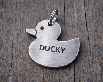 May include: A brushed metal pet tag in the shape of a duck. The tag has a ring for attachment and the name "DUCKY" engraved in a bold font. The background is a weathered wooden surface.