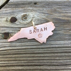May include: A copper-colored North Carolina state-shaped dog tag with the name "SARAH" engraved on it and a paw print below the name.