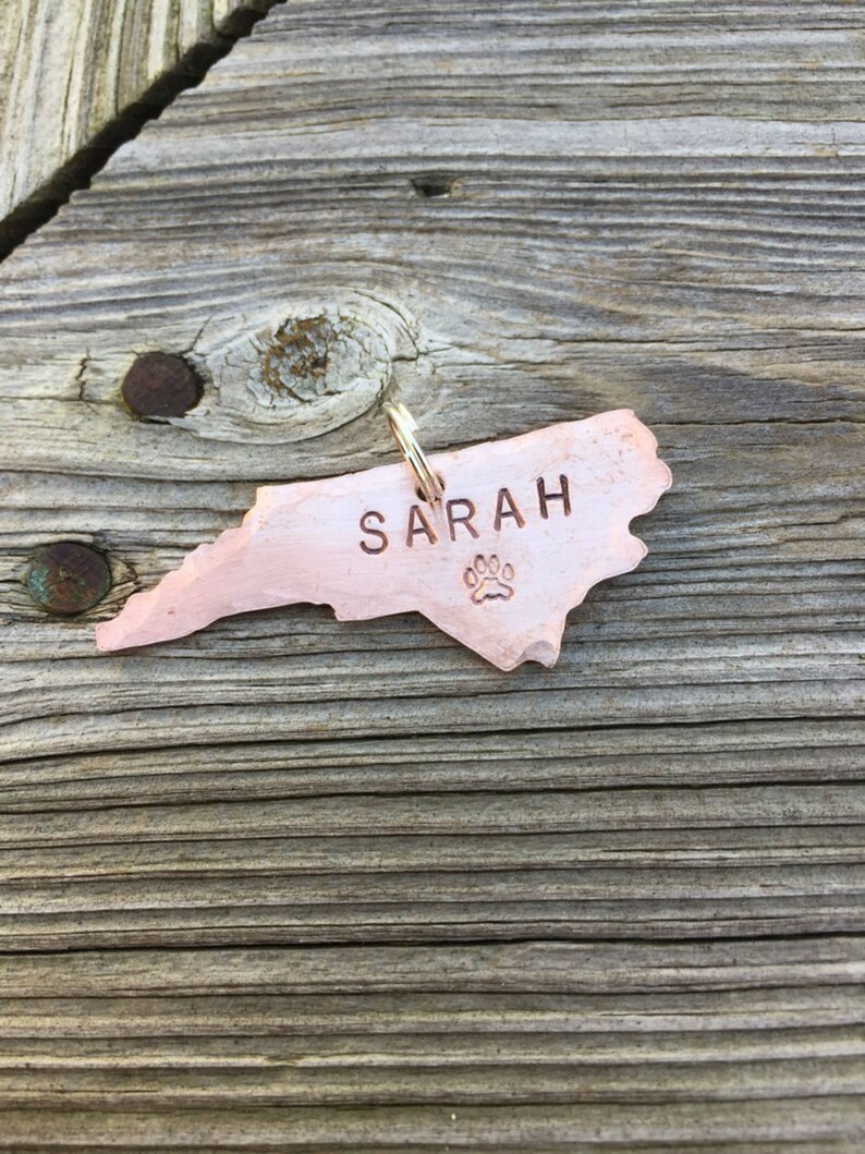 May include: A copper-colored North Carolina state-shaped dog tag with the name "SARAH" engraved on it and a paw print below the name.