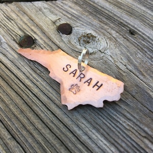 May include: A copper dog tag in the shape of North Carolina with the name "SARAH" engraved on it and a paw print design.