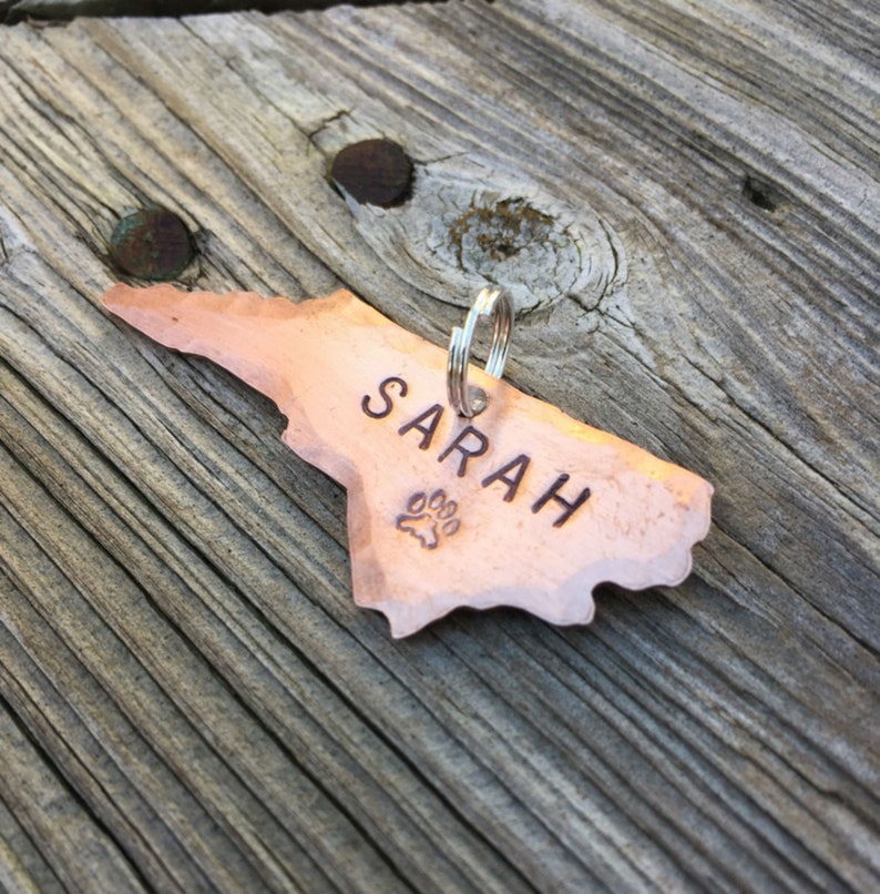 May include: A copper dog tag in the shape of North Carolina with the name "SARAH" engraved on it and a paw print design.