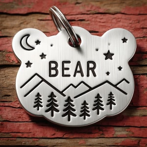 May include: Silver bear-shaped dog tag with a mountain and forest design, a crescent moon, stars, and the word "BEAR" engraved on it.