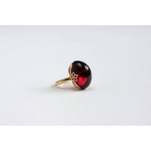 May include: A gold ring with a large, oval-shaped, dark red gemstone. The ring has a delicate floral design on the band.