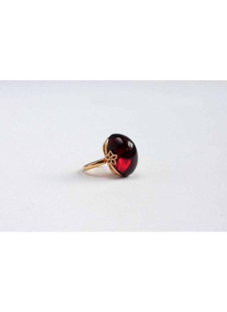 May include: A gold ring with a large, oval-shaped, dark red gemstone. The ring has a delicate floral design on the band.
