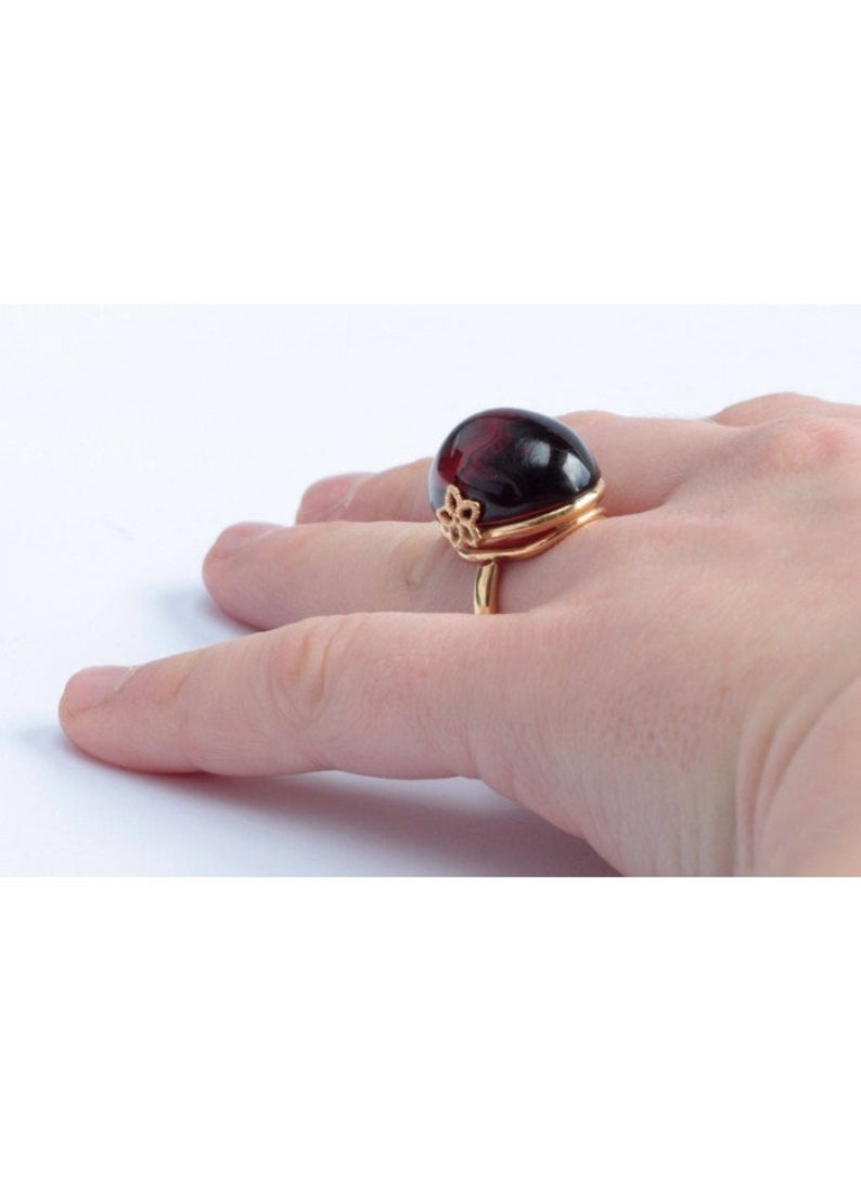 May include: A gold ring with a large, dark red oval gemstone. The ring has a delicate floral design on the band.