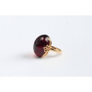 May include: A gold ring with a large, oval-shaped, dark red gemstone. The ring has a delicate floral design on the band.