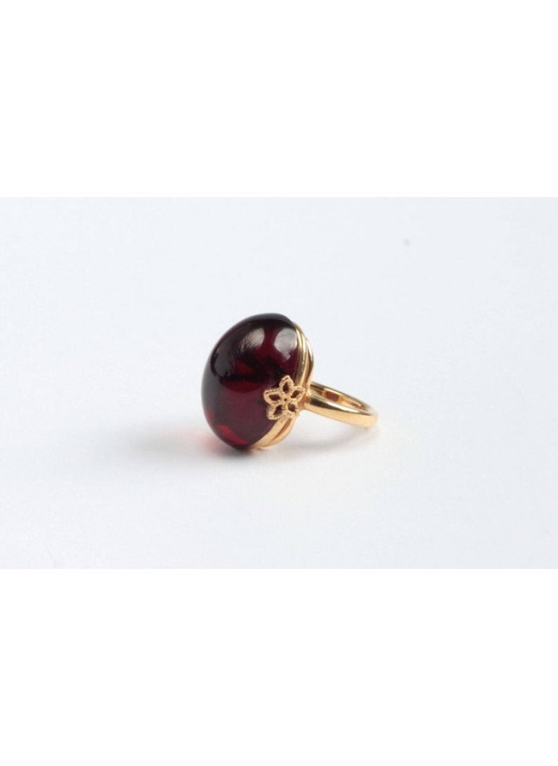 May include: A gold ring with a large, oval-shaped, dark red gemstone. The ring has a delicate floral design on the band.