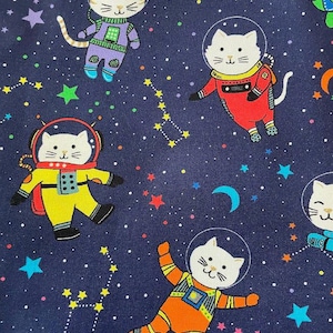 May include: A navy blue fabric featuring cartoon cats in astronaut suits floating among stars, constellations, and crescent moons. The cats wear colorful space suits in yellow, orange, red, and purple. The background is filled with colorful stars.