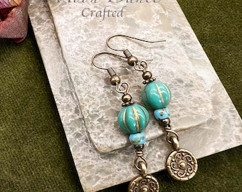 Può includere: Orecchini fatti a mano con dettagli turchese e oro. Gli orecchini presentano perline turchesi, piccole perline blu e ciondoli color oro. Il testo "Moon Dance Crafted" è visibile sullo sfondo.