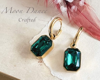 Può includere: Un paio di orecchini a cerchio dorati con cristalli color smeraldo rettangolari sfaccettati. Il testo Moon Dance Crafted è visibile nell'angolo in alto a sinistra. Gli orecchini sono mostrati accanto a una piuma rossa a pois bianchi.