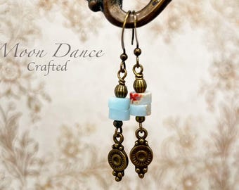 Può includere: Orecchini fatti a mano dall'estetica vintage. Ogni orecchino presenta una perla azzurra, una perla fantasia e un ciondolo decorativo color bronzo. Gli orecchini sono sospesi da ganci per orecchie color bronzo. Il testo "Moon Dance Crafted" è visibile.