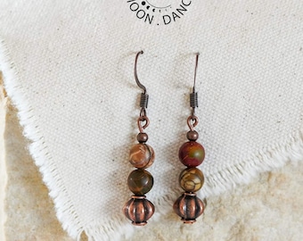 Può includere: Un paio di orecchini pendenti con ganci e componenti color rame. Ogni orecchino è composto da due perline rotonde in pietra dai toni della terra e una perlina in metallo striato color rame alla base. Il testo MOON DANCE è visibile sopra.