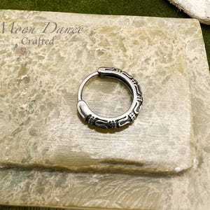 May include: A silver hoop earring with an ornate, engraved design. The earring is circular with a hinged closure. The text "Moon Dance Crafted" is visible in the background. The earring is displayed on a light green, textured surface.