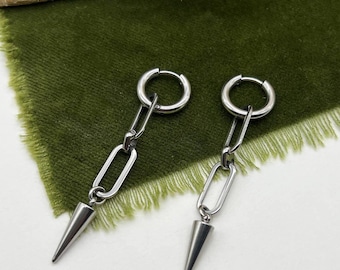 Può includere: Un paio di orecchini color argento con un design a catena e un pendente a forma di cono. Gli orecchini sono presentati su un tessuto verde, con il testo "Moon Dance" in alto a destra.