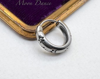 Può includere: Un orecchino a cerchio argentato con intagli a forma di stella. L'orecchino è su una superficie bianca, con uno sfondo viola incorniciato d'oro nell'angolo in alto a sinistra. Il testo "Moon Dance" è visibile.