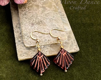 Può includere: Un paio di orecchini color oro con pendenti a forma di ventaglio rosso scuro. Gli orecchini sono esposti su una superficie di pietra grigio chiaro, con il testo "Moon Dance Crafted" visibile. Lo sfondo è un tessuto verde scuro.