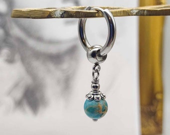 May include: A silver hoop earring with a blue and brown marbled stone charm hanging from it. The earring is on a gold surface.
