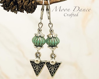 Può includere: Un paio di orecchini pendenti color argento con una perla scanalata color turchese, decorata con tappi in filigrana d'argento e un ciondolo triangolare con dettagli neri incisi. Il testo Moon Dance Crafted è visibile in alto a destra.