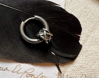 Può includere: Un orecchino a cerchio argentato con un design intrecciato, appoggiato su una piuma nera. L'orecchino presenta un piccolo e intricato dettaglio a forma di nodo al centro. Lo sfondo include un pezzo di carta con testo manoscritto.