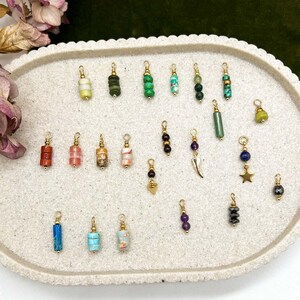 Peut inclure: Une collection de breloques en pierres précieuses avec des attaches dorées, présentées sur un plateau ovale clair. Les breloques présentent une variété de couleurs et de textures, notamment du vert, du bleu, du rouge et du violet. Les breloques sont petites et délicates.