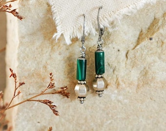 Può includere: Un paio di orecchini pendenti con perline cilindriche in malachite verde e perline metalliche rotonde sospese a ganci color argento. Gli orecchini sono esposti su una superficie di pietra ruvida con un pezzo di tessuto.