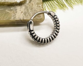 Può includere: Un orecchino a cerchio color argento con un design strutturato. L'orecchino è circolare e presenta una serie di linee verticali lungo il bordo esterno. Il testo "Moon Dance" è scritto in corsivo nella parte inferiore dell'immagine.