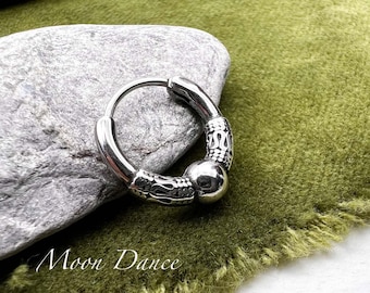 May include: A silver hoop earring with ornate detailing. The earring features a decorative design on the thicker portion and a smooth, rounded ball. The earring is displayed on a gray stone and green fabric. The text "Moon Dance" is visible.