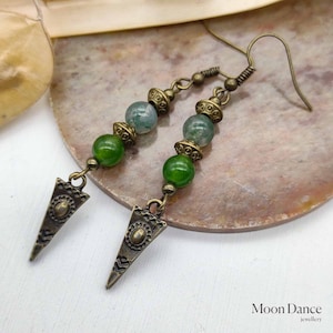 May include: A pair of green agate gemstone earrings with bronze triangle charms and bronze beads. The earrings are on a light brown surface.