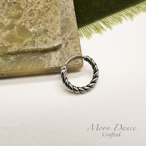 May include: A silver-coloured, twisted hoop earring. The earring features a braided rope design and a hinged closure. The earring is on a white surface with the text "Moon Dance Crafted" in the bottom right corner.