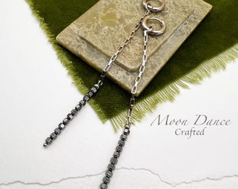 Può includere: Un paio di lunghi orecchini a catena d'argento con accenti di perline nere. Gli orecchini sono appesi su una superficie marrone chiaro con il testo "Moon Dance Crafted" scritto in nero.
