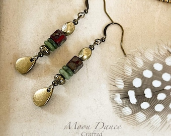 Può includere: Orecchini pendenti con ganci in ottone anticato, piccole perline in ottone testurizzato, perline quadrate screziate rosso e marrone, perline a disco verdi e un ciondolo a goccia in ottone. Il testo Moon Dance Crafted è visibile in basso.