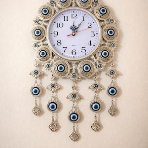 Op de afbeelding: Een decoratieve wandklok met een evil eye-ontwerp. De klok heeft een witte wijzerplaat met zwarte cijfers en wijzers, omgeven door een goudkleurig frame versierd met blauwe en witte evil eye-bedels. Er zijn ook bungelende bedels met evil eye-hangers.