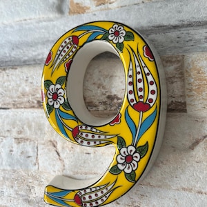 May include: A yellow ceramic number nine with a white background and a floral design in red, white, blue, and green. The number is a decorative piece for a home.