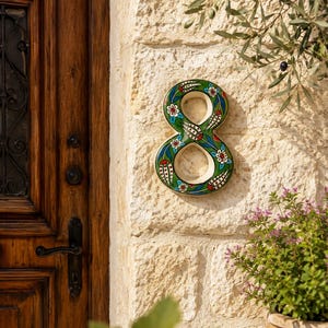 May include: A ceramic house number eight featuring a green background with hand-painted floral patterns in red, white, and blue. The number has a glossy finish and is mounted on a textured stone wall next to a wooden door and a potted plant.