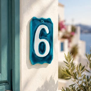 Peut inclure: Plaque de numéro de maison en céramique turquoise avec le chiffre 6 blanc. Fixée sur un mur blanc texturé. Le fond montre une porte bleue et une vue floue d'une scène côtière.