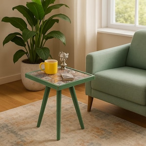 May include: A small, square side table with a green frame and legs. The tabletop features a mosaic tile design with various colors. A yellow mug, a small book, and a silver sculpture sit on the table. A green armchair and a potted plant are in the background.