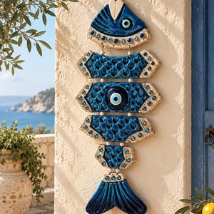 May include: A decorative wall hanging shaped like a fish, constructed from five interconnected ceramic segments in deep blue and cream. The design features multiple small circular evil eye beads and a central larger evil eye bead on the body.