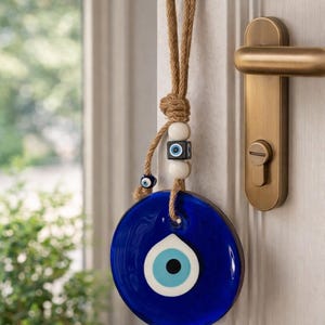 Op de afbeelding: Een cirkelvormig wandornament van blauw glas met een oogmotief, opgehangen aan een dik touw van jute. Het ornament heeft een centraal wit en lichtblauw oogontwerp, geaccentueerd met witte bolvormige kralen en een kleine vierkante kraal met oogmotief.
