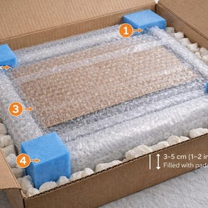 May include: A brown cardboard shipping box containing a rectangular item wrapped in clear bubble wrap. Four blue foam corner protectors are placed at the corners. White packing peanuts fill the gaps. Text indicates 3-5 cm (1-2 inch) padding.