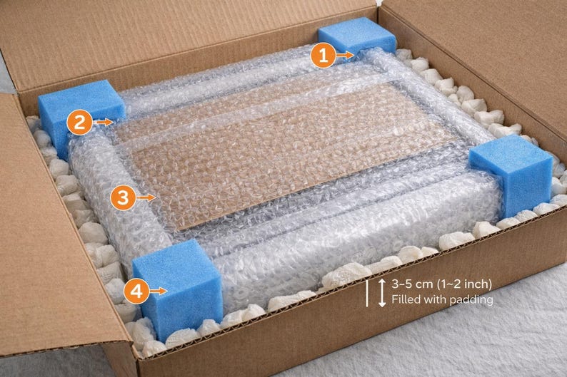 May include: A brown cardboard shipping box containing a rectangular item wrapped in clear bubble wrap. Four blue foam corner protectors are placed at the corners. White packing peanuts fill the gaps. Text indicates 3-5 cm (1-2 inch) padding.