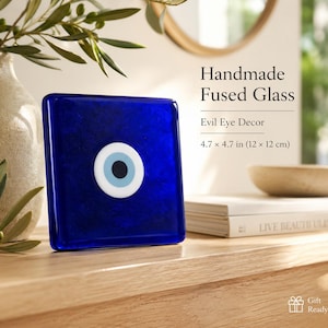 May include: A square handmade fused glass piece featuring a deep blue background with a centred evil eye motif consisting of white, light blue, and black circles. The text reads Handmade Fused Glass, Evil Eye Decor, 4.7 x 4.7 in (12 x 12 cm).