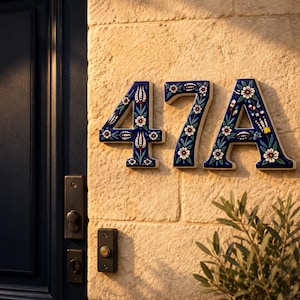 May include: Ceramic house number plaque displaying the characters 47A in a dark blue color with a white, red, and green floral pattern. The numbers and letter are mounted on a light-colored stone wall next to a dark blue door and a doorbell.