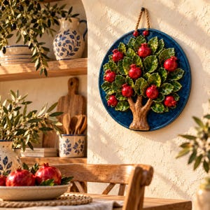 May include: A circular ceramic wall plaque featuring a relief sculpture of a tree with a brown trunk, green leaves, and bright red pomegranates against a deep blue background. The plaque is suspended by a thick, twisted rope cord at the top.