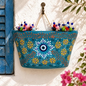 May include: A teal ceramic wall planter featuring a central blue and white evil eye motif surrounded by hand-painted yellow flowers and orange vines. The planter is suspended by a jute rope from a metal hook and decorated with colourful beads.
