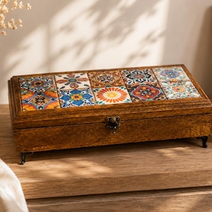 May include: A rectangular wooden storage box featuring a hinged lid decorated with five colorful square tiles displaying intricate floral and geometric patterns. The box has a dark brown finish, a metal latch, and four small black decorative feet.