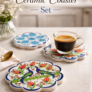 Può includere: Set di sottobicchieri in ceramica fatti a mano. I sottobicchieri presentano motivi floreali e di uccelli blu, rossi e verdi su una base bianca. Un sottobicchiere contiene una tazza di caffè. Il testo "Handmade Ceramic Coaster - Set" è in alto.