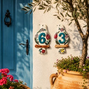 Pode incluir: Duas placas de número de casa em cerâmica com formato de pássaro, exibindo os números 6 e 3 em relevo na cor azul-petróleo. As placas brancas são decoradas com motivos florais pintados à mão em vermelho, rosa, verde e amarelo na parede.